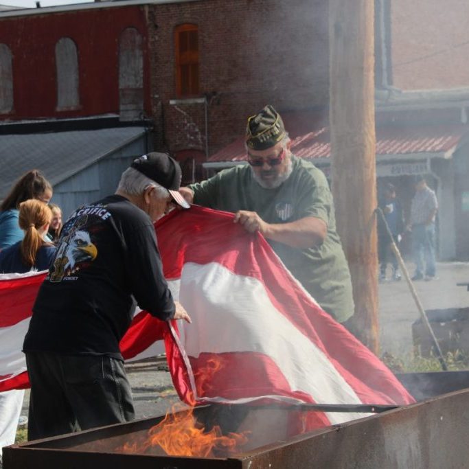 Photo by Katey Wesley
Veterans Ed Folger, left, and Aaron Hall lower a portion of the large flag into the fire. Not pictured are the students who are supporting the other end of the flag as they bring it to the fire, treating the flag with respect even during its final moments.